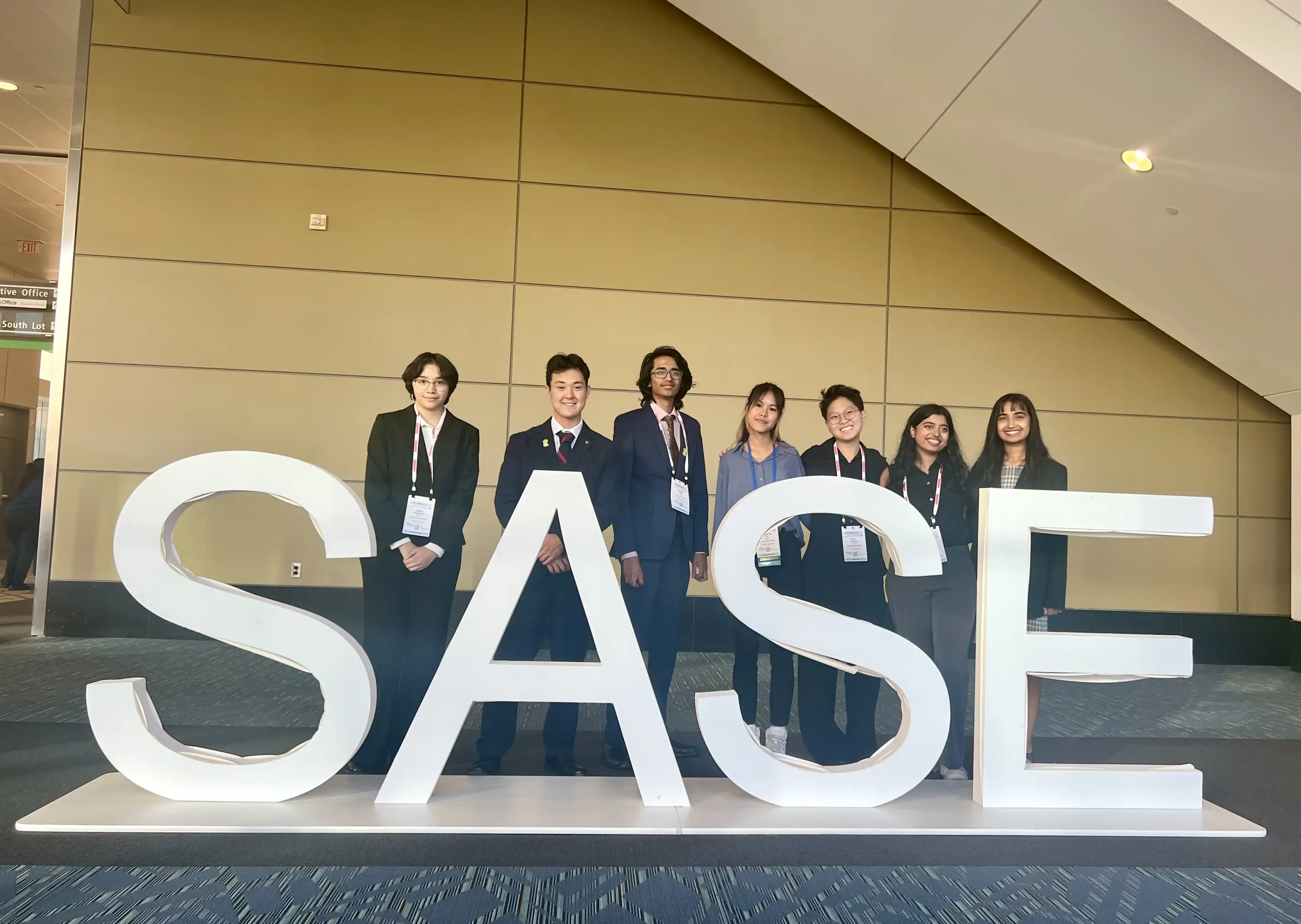 Zephyr, Mason, Chinmay, Su, Aro, Stef, and Anisha take a photo in front of a large SASE logo sign in Boston, MA